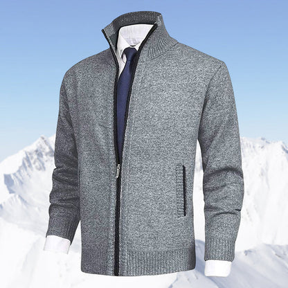 Grauer Herren-Strickjacke mit Reißverschluss, kombiniert mit weißem Hemd und blauer Krawatte, vor schneebedeckten Bergen. Elegante Wintermode.