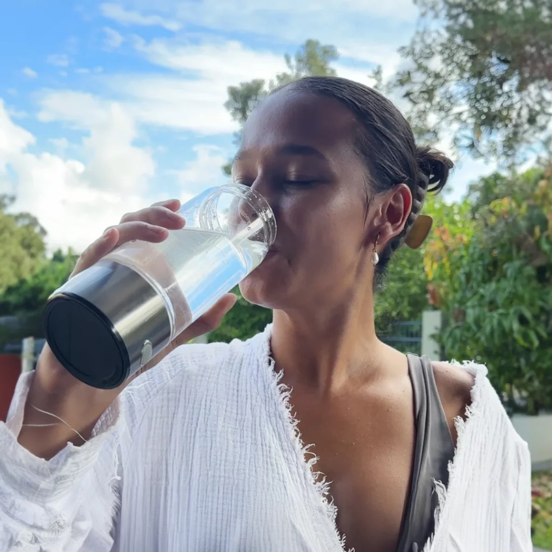 Frau trinkt Wasser aus Glas, Sommerkleidung, draußen, blauer Himmel, gesunde Lebensweise, Hydration, Naturhintergrund, Erfrischung.