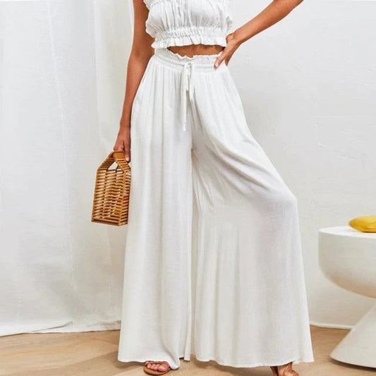 Frau in weißem, ärmellosem Sommeroutfit mit weitem Hosenrock, hält eine geflochtene Handtasche. Elegante, luftige Mode für warme Tage.