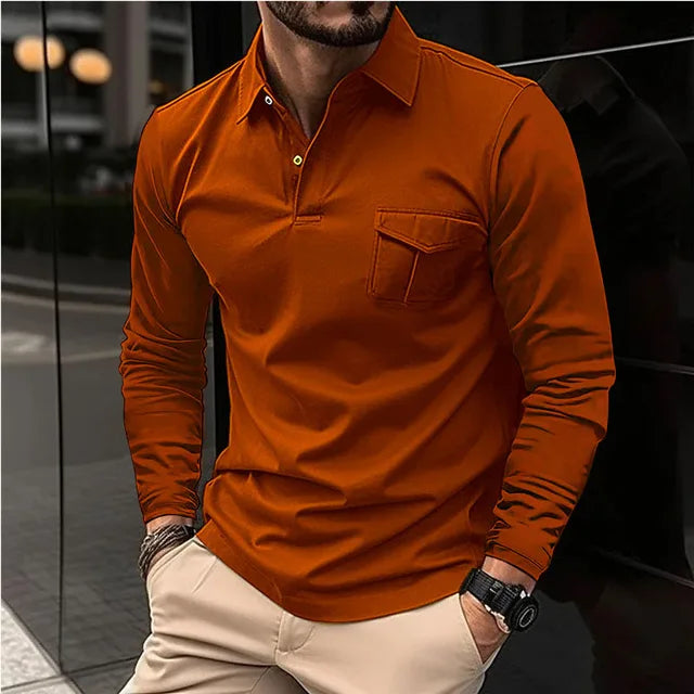 Mann in orangefarbenem Langarmhemd mit Brusttasche, beiger Hose und Armbanduhr, steht vor einem modernen Gebäude. Elegante Herrenmode, urbaner Stil.