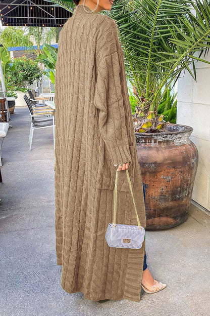 Frau in langem, beigem Strickmantel, hält kleine Handtasche. Steht draußen neben großen Pflanzen. Modetrend, Herbstmode, Strickkleidung.
