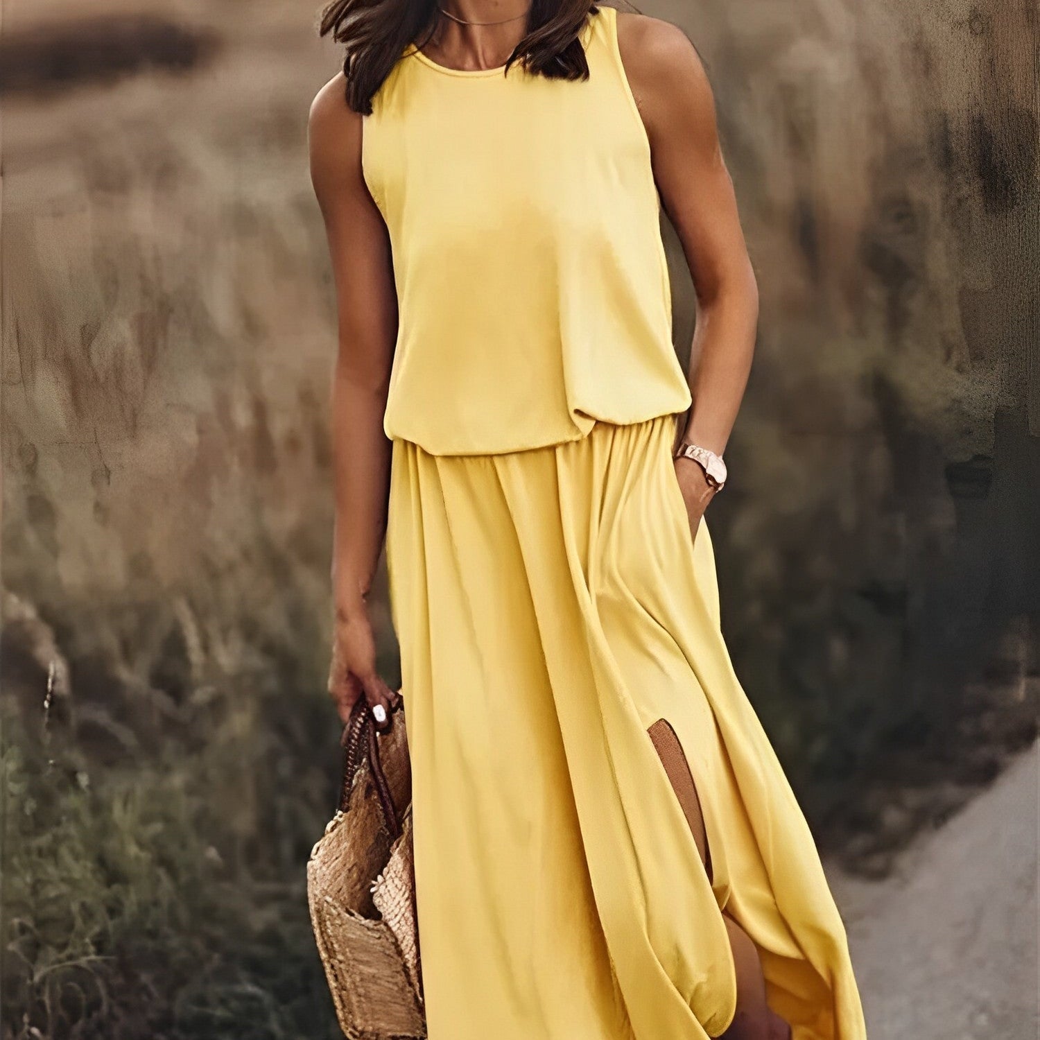 Frau in gelbem Sommerkleid mit Schlitz, hält Strohtasche, steht auf Feldweg. Modetrend, Sommermode, elegantes Damenoutfit, Freizeitkleidung.