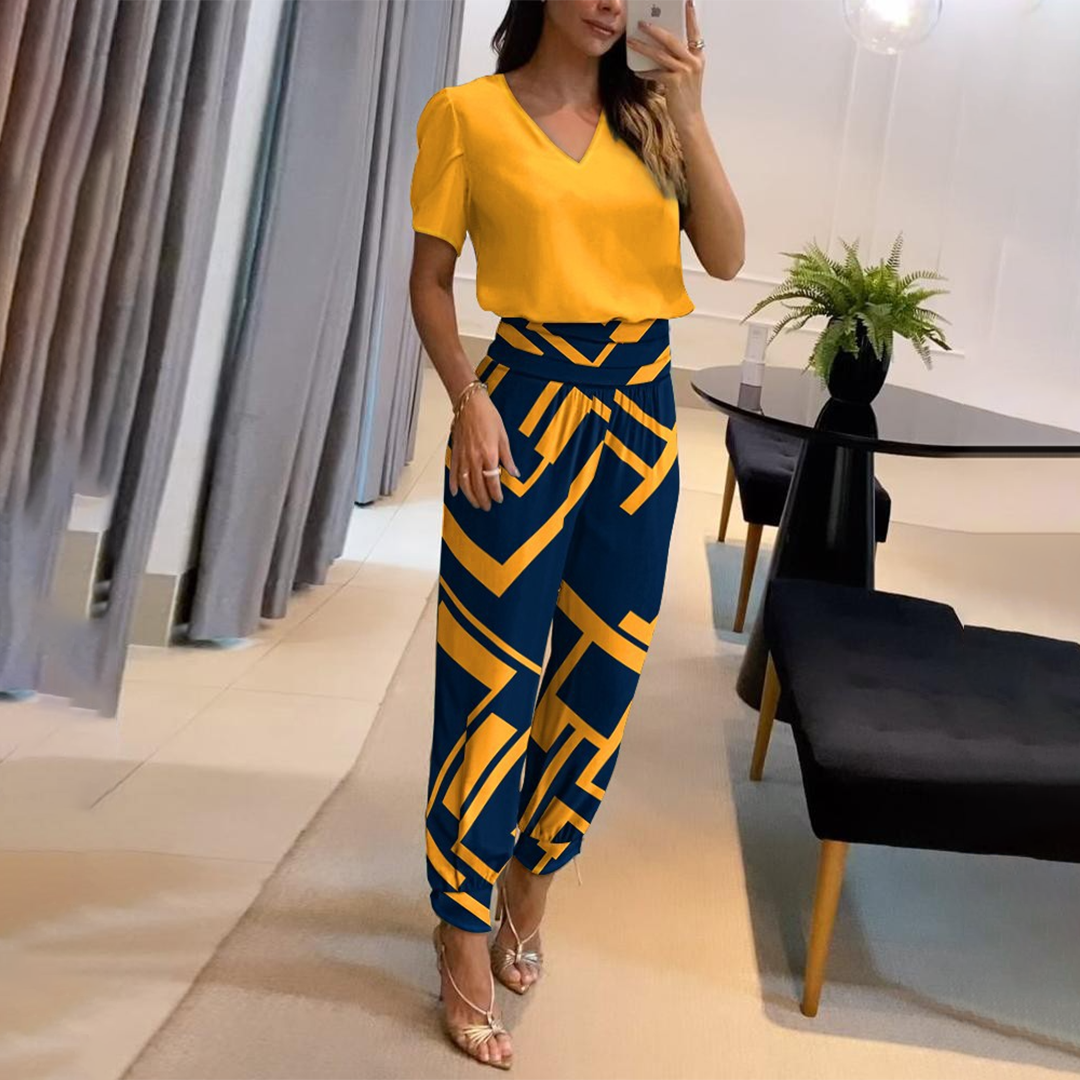 Frau in gelbem V-Ausschnitt-Oberteil und geometrisch gemusterter blauer Hose, steht in modernem Wohnzimmer mit Pflanzen und Spiegel, modisch elegant.