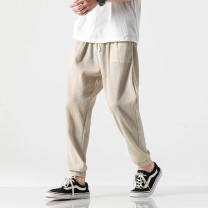 Beige Herren-Jogginghose aus Leinen, kombiniert mit schwarzen Sneakern und weißem T-Shirt. Bequeme Freizeitmode für Männer.