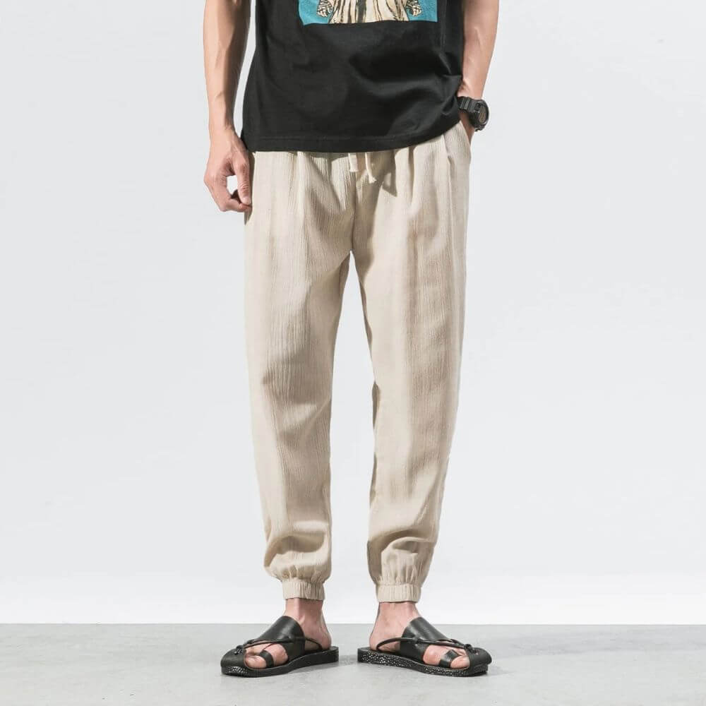 Mann trägt beige Leinenhose und schwarze Sandalen, lässiger Stil, Sommermode, Herrenbekleidung, stehend vor weißem Hintergrund.