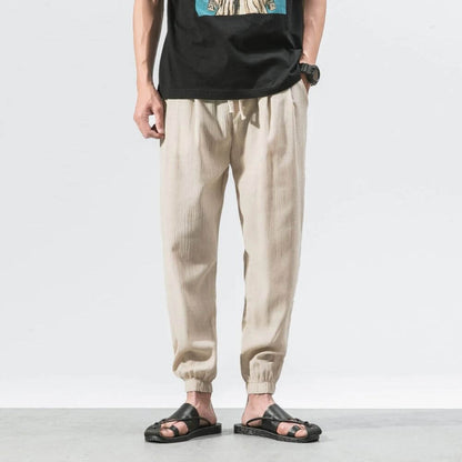 Mann trägt beige Leinenhose und schwarze Sandalen, lässiger Stil, Sommermode, Herrenbekleidung, stehend vor weißem Hintergrund.