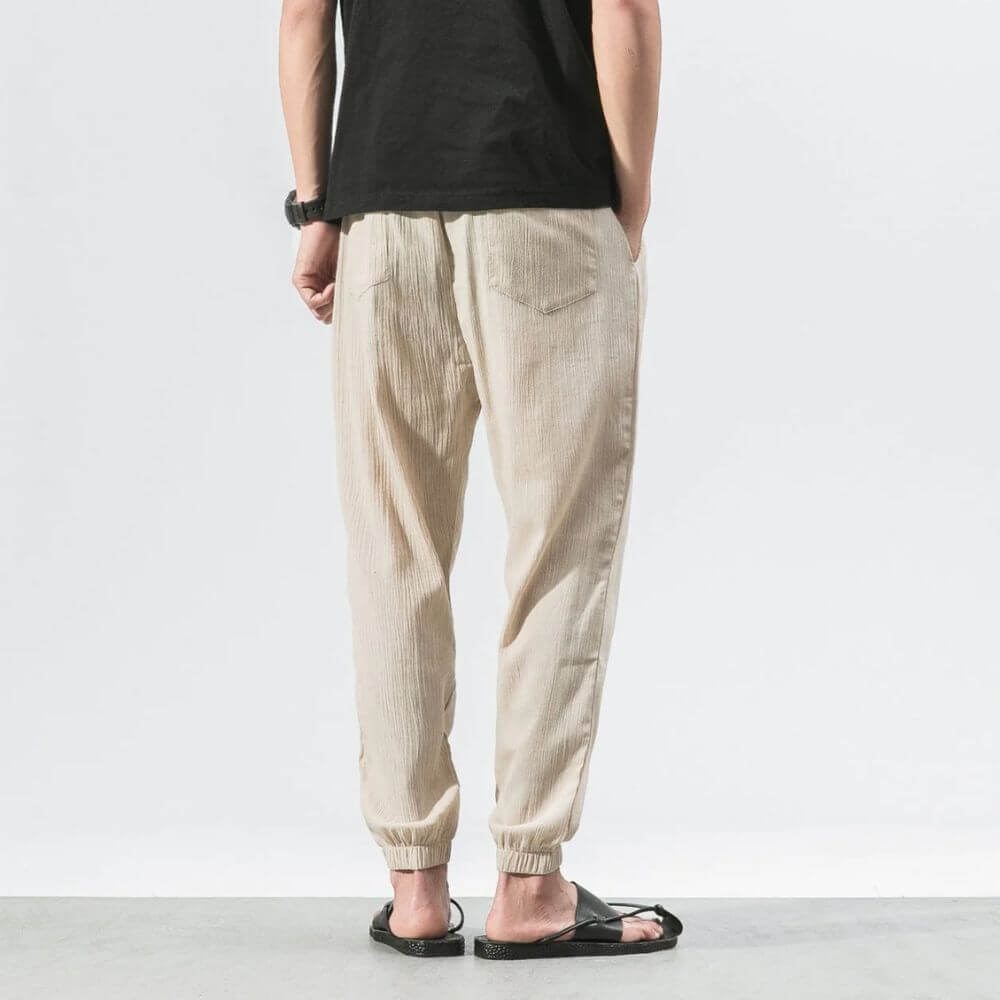 Rückansicht einer Person in beigen Leinenhosen und schwarzem T-Shirt, stehend auf grauem Boden. Lässige Sommermode, Herrenbekleidung, Freizeitlook.