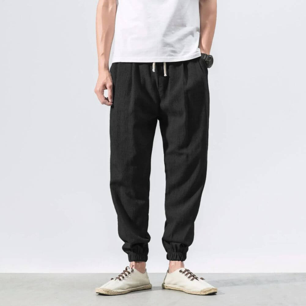 Mann trägt schwarze Leinenhose und weiße Sneakers vor grauem Hintergrund. Modische Herrenbekleidung, lässiger Stil, komfortable Passform.