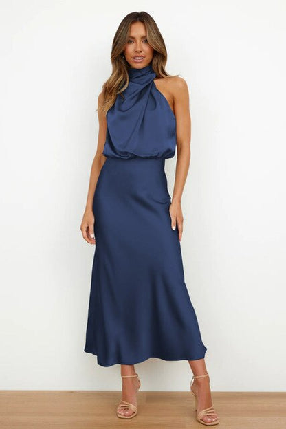 Frau in elegantem, blauem Satinkleid mit hohem Kragen und ärmellosem Design, stehend vor weißem Hintergrund, trägt beige High Heels.