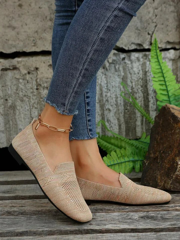 Damenbeine in blauen Jeans mit beige Loafern auf Holzboden, umgeben von grünen Farnen. Modische Schuhe, Freizeitlook, Outdoor.