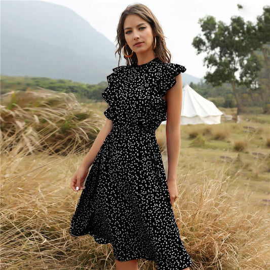 Frau in schwarzem, gepunktetem Kleid steht in natürlicher Landschaft mit Zelt im Hintergrund. Sommermode, Outdoor, stilvoll, elegant.