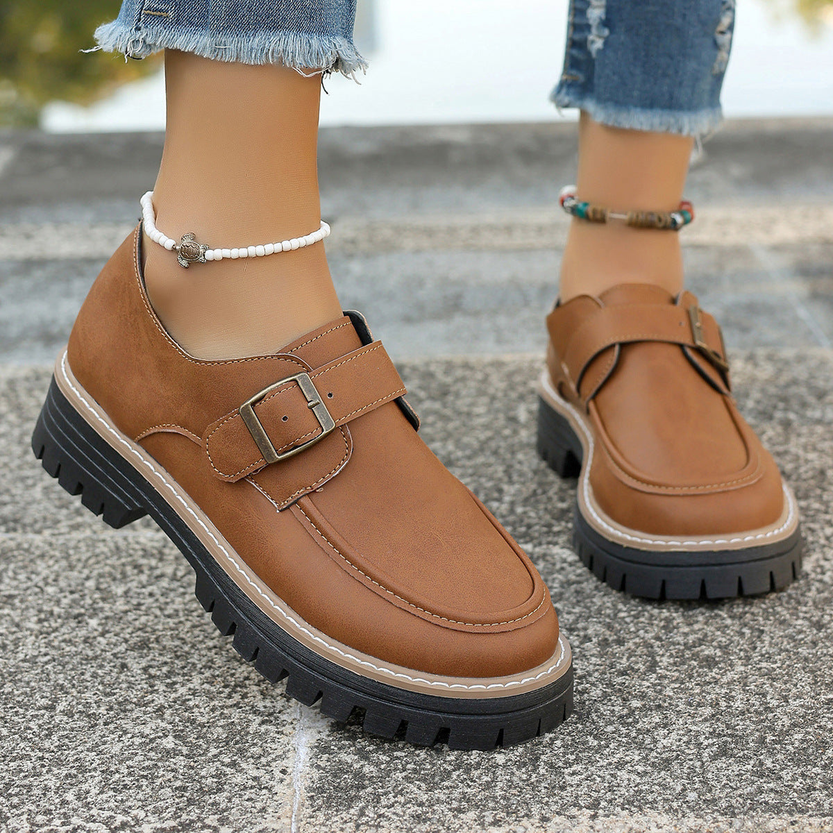 Braune Damen-Loafer mit Schnalle, dicker Sohle und Ziernähten, getragen mit Jeans und Fußkettchen. Modische Herbstschuhe für Damen.