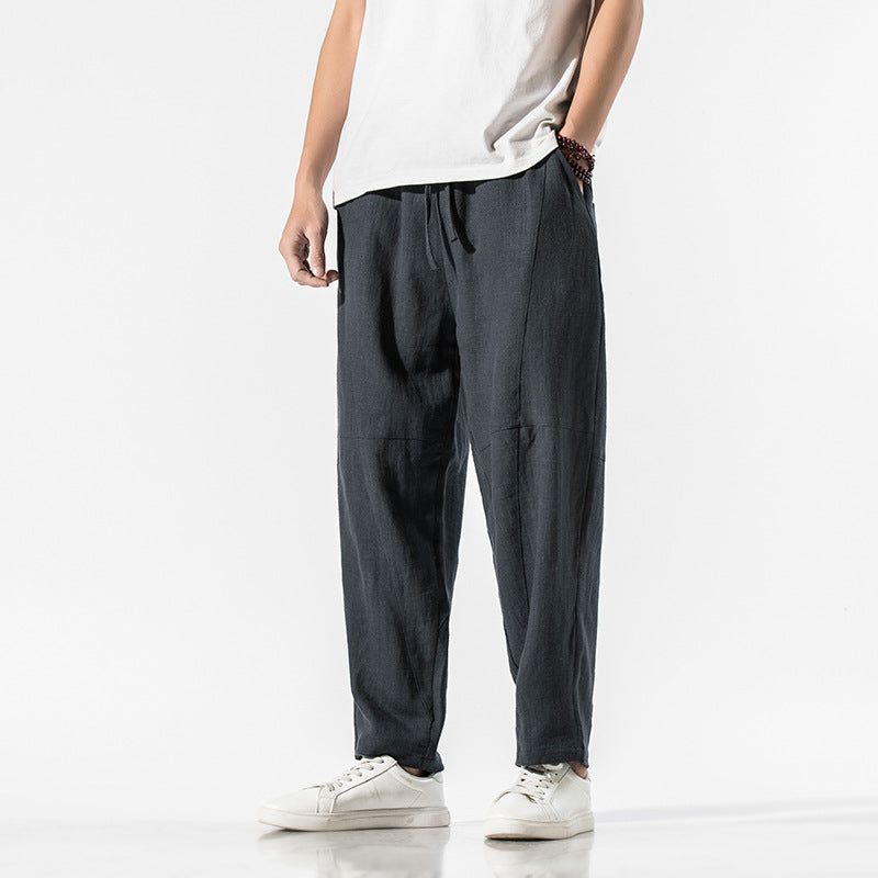 Mann in lässiger, dunkelgrauer Leinenhose und weißem T-Shirt, trägt weiße Sneaker. Moderne Herrenmode, bequeme Freizeitkleidung.
