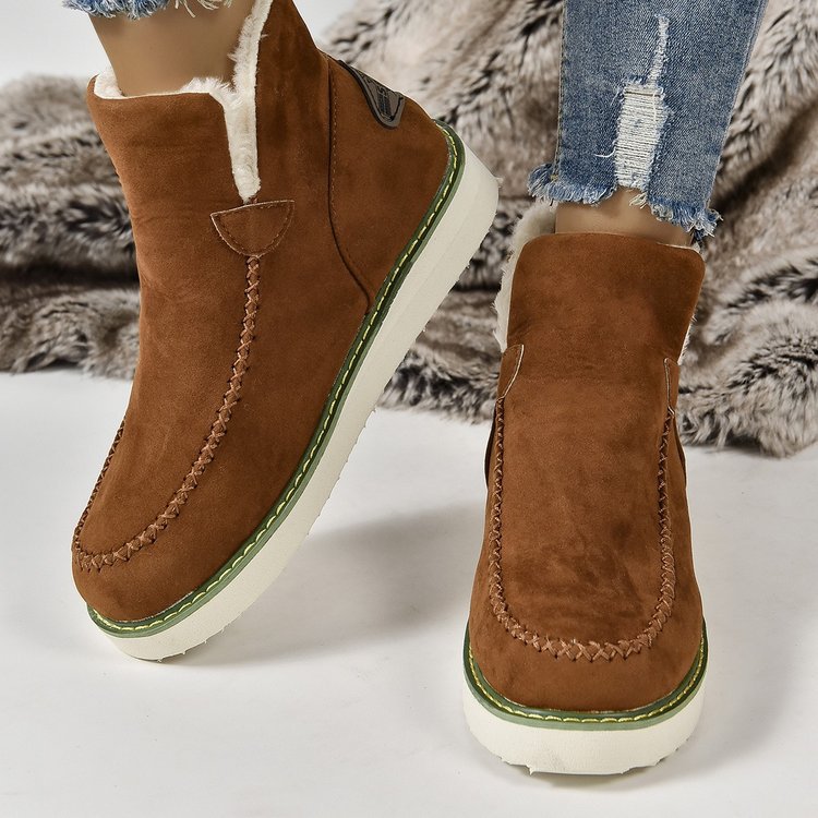 Braune Wildleder-Winterstiefel mit weißer Sohle und grüner Ziernaht, getragen mit zerrissenen Jeans auf einem Fellteppich.