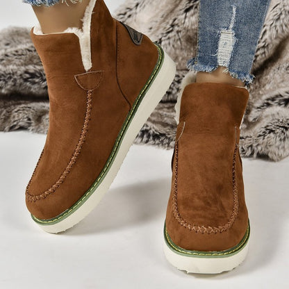 Braune Wildleder-Winterstiefel mit weißer Sohle und grüner Ziernaht, getragen mit zerrissenen Jeans auf einem Fellteppich.