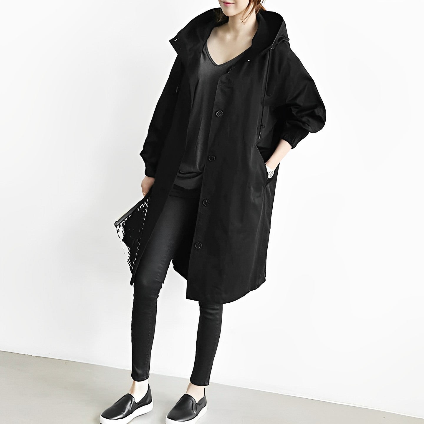 Frau in schwarzem Oversize-Mantel, schwarzer Hose und Slip-On-Schuhen, hält eine Clutch. Modetrend, lässiger Look, Streetstyle.
