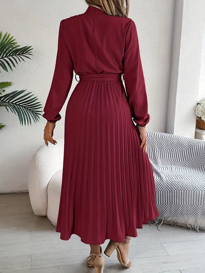 Frau in elegantem, langem, weinrotem Kleid mit Faltenrock, steht in modernem Wohnzimmer. Stilvolle Mode, Damenbekleidung, Herbsttrends.