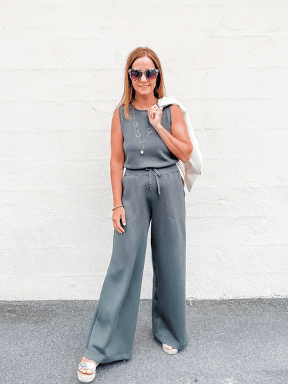 Frau in grauem Jumpsuit vor weißer Wand, trägt Sonnenbrille und Sandalen, hält weiße Jacke. Modetrend, Sommeroutfit, lässiger Stil.