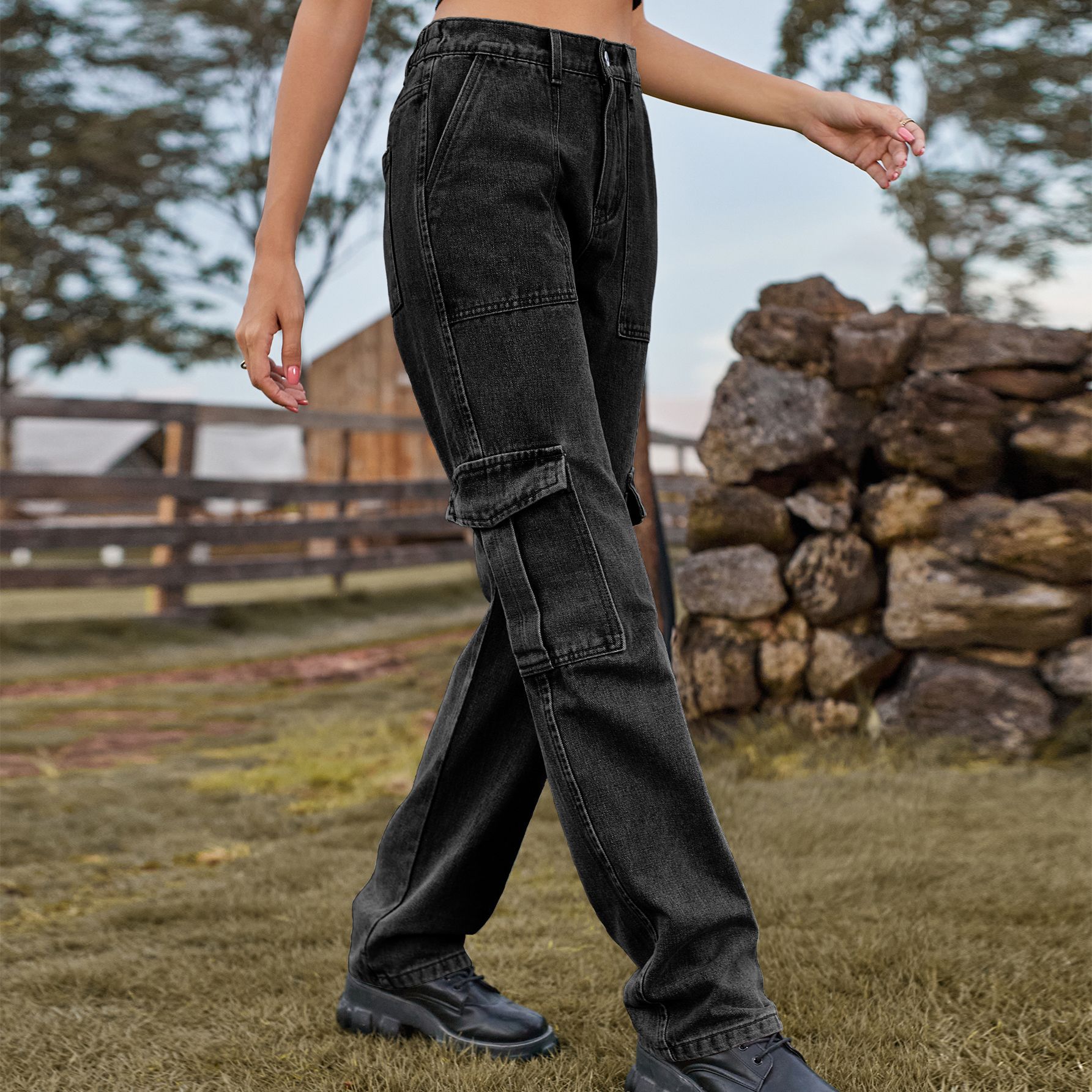 Frau in schwarzer Cargohose spaziert auf einem Bauernhof, im Hintergrund Holzzaun und Steinhaufen. Modetrend, Outdoor, Freizeitkleidung.