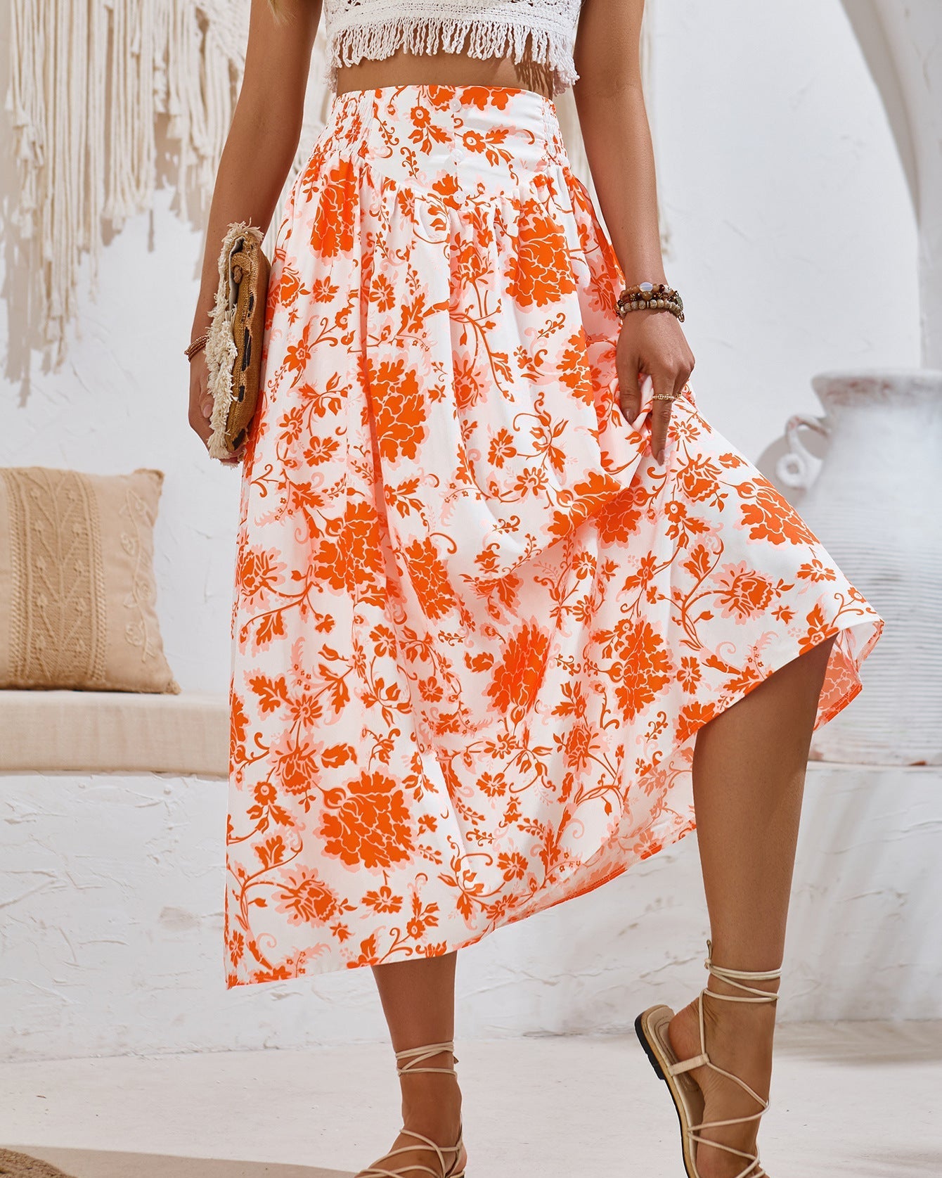 Frau in weißem, ärmellosem Oberteil und orange-weißem Blumenrock, hält eine Tasche. Sommermode, Boho-Stil, modische Sandalen.