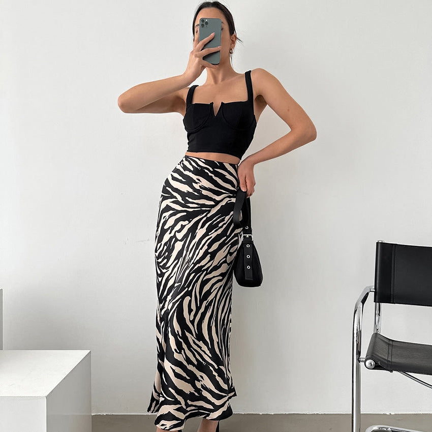 Frau in schwarzem Top und Zebra-Muster-Rock, hält Smartphone, posiert vor weißer Wand. Modetrend, stilvolle Kleidung, modernes Outfit.