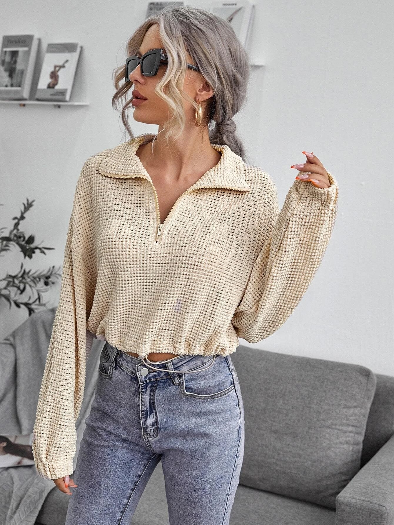 Frau mit blonden Haaren trägt beige Strickpullover und Jeans, steht in modernem Wohnzimmer. Stilvolle Mode, lässiger Look, Sonnenbrille.