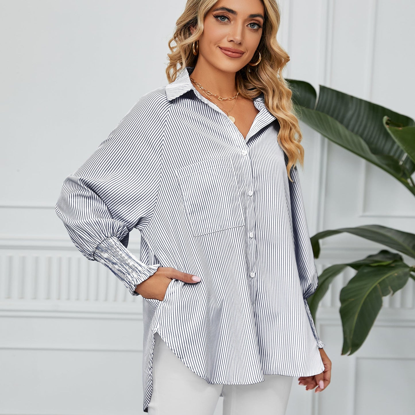 Blonde Frau in gestreifter Bluse, lächelt, steht vor weißer Wand mit Pflanze. Modetrend, Damenmode, elegante Freizeitkleidung, stilvoll.