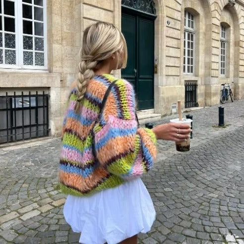 Frau mit geflochtenem Haar trägt bunten Strickpullover und hält Bubble Tea, spaziert auf Kopfsteinpflasterstraße vor historischen Gebäuden.