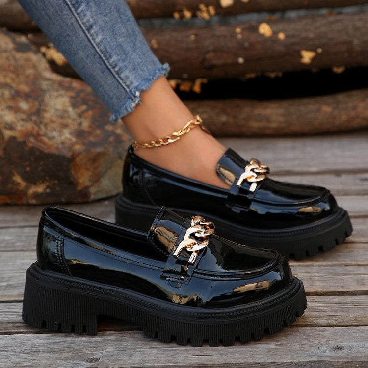 Schwarze Lack-Loafer mit goldener Kette und dicker Sohle, getragen von Person in Jeans. Modische Damenschuhe, trendiger Look, Herbstmode.