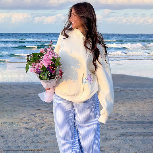 Frau am Strand mit Blumenstrauß, trägt weißen Hoodie und gestreifte Hose, lächelt vor Meereswellen. Entspannter Strandspaziergang bei Sonnenuntergang.