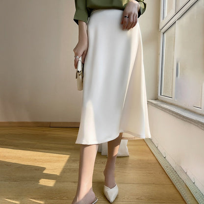 Eleganter weißer Midirock aus Satin, kombiniert mit grüner Bluse und weißen Pumps, steht auf hellem Holzboden vor einem Fenster.
