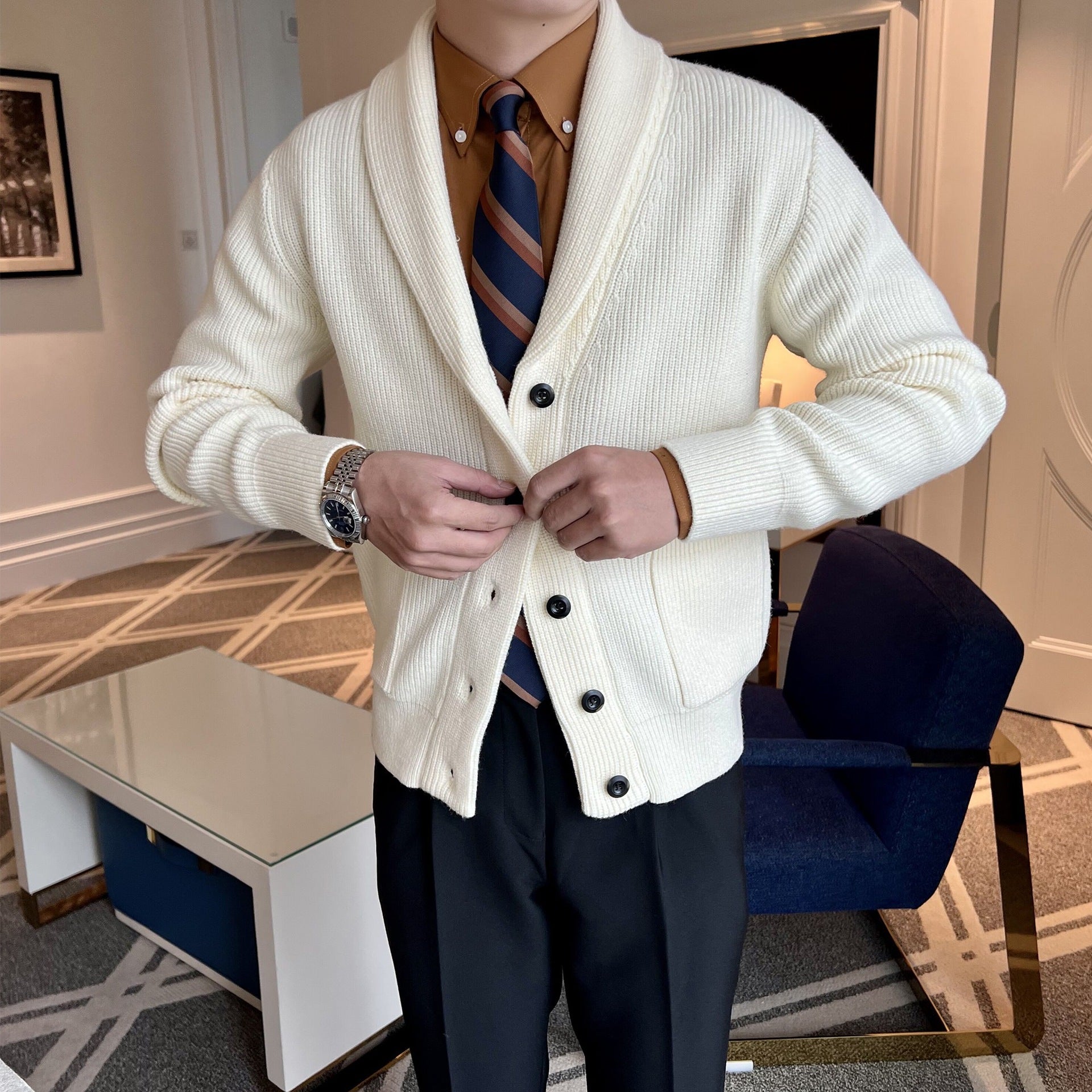 Mann in cremefarbener Strickjacke mit Knöpfen, braunem Hemd und gestreifter Krawatte in modernem Wohnzimmer. Elegante Herrenmode, stilvoll gekleidet.