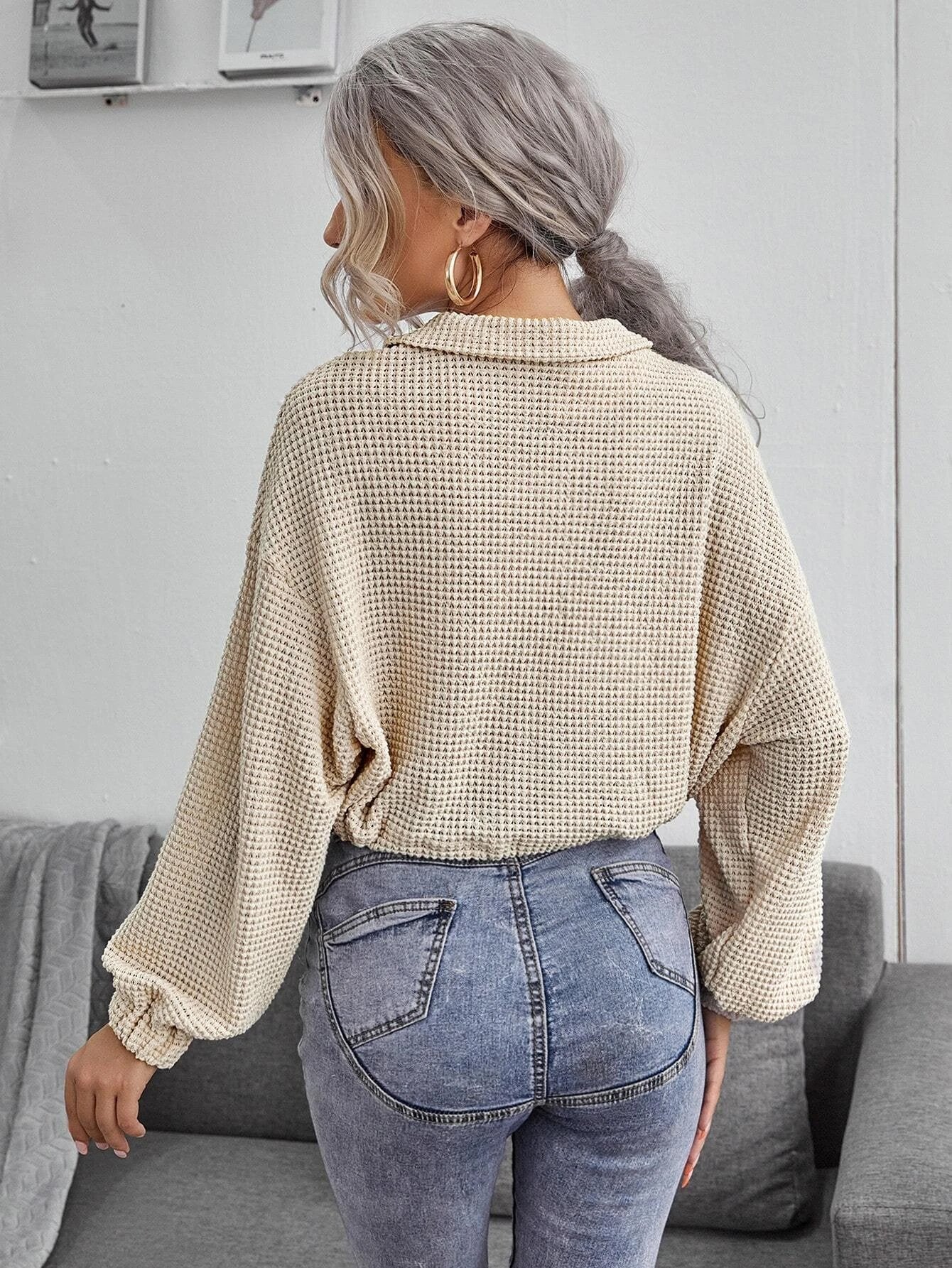 Frau mit grauem Haar trägt beige Strickpullover und blaue Jeans, steht in modernem Wohnzimmer. Modetrend, lässiger Stil, Herbstmode.