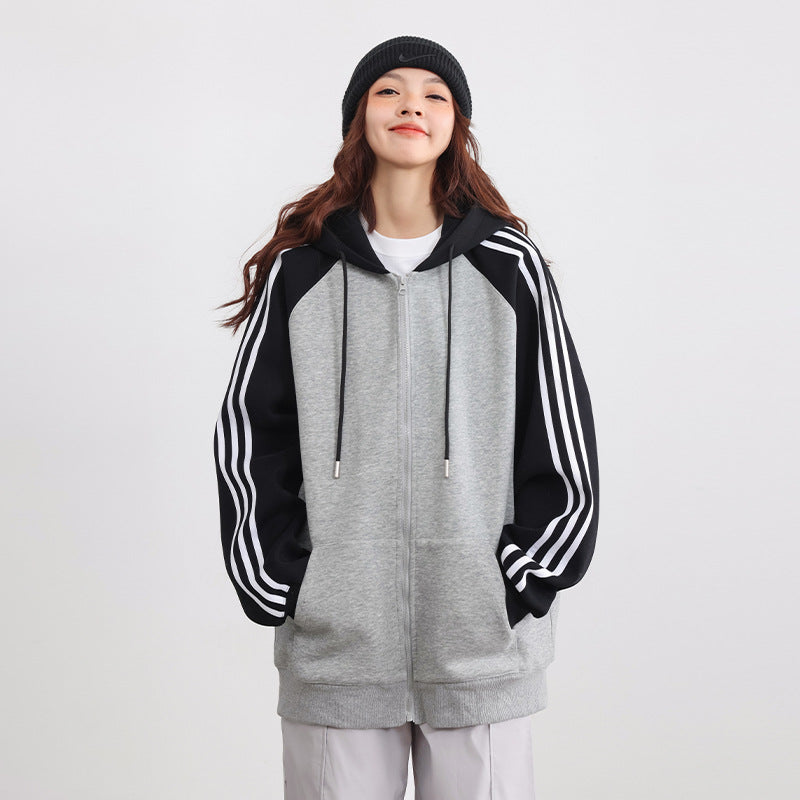 Frau in grauem Hoodie mit schwarzen Ärmeln und weißen Streifen, trägt schwarze Mütze. Lächelnd, lässiger Stil, modische Freizeitkleidung.