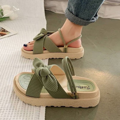 Grüne Sandalen mit Schleifen, flache Sohle und Riemen, getragen von einer Person auf grauem Boden. Modische Sommerschuhe für Damen.