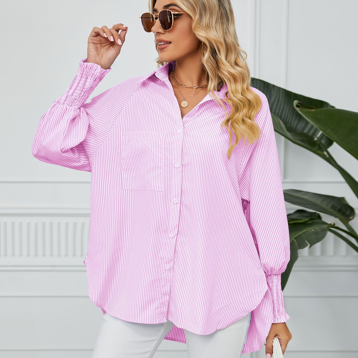 Blonde Frau in rosa gestreifter Bluse und Sonnenbrille, posiert vor weißer Wand mit grüner Pflanze. Modetrend, Damenmode, lässiger Stil.