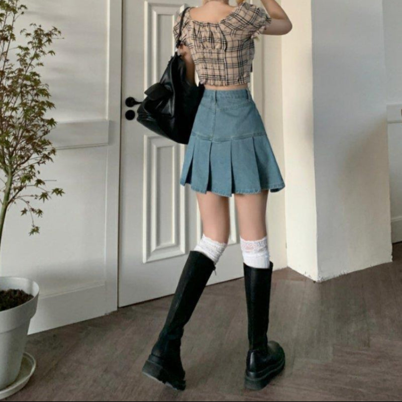 Junge Frau in kariertem Top und Jeansrock, trägt hohe schwarze Stiefel und weiße Kniestrümpfe, steht vor einer weißen Tür, modisch, urbaner Stil.