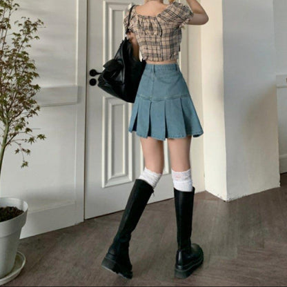 Junge Frau in kariertem Top und Jeansrock, trägt hohe schwarze Stiefel und weiße Kniestrümpfe, steht vor einer weißen Tür, modisch, urbaner Stil.