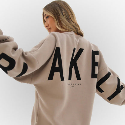 Frau in beigem Oversize-Pullover mit großem schwarzen Schriftzug "AKELY" auf Rücken und Ärmeln, modisches Streetwear-Outfit, seitliches Profil.