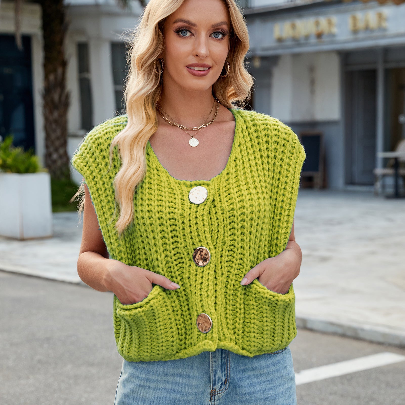 Blonde Frau trägt gelbe Strickweste mit großen Knöpfen und Taschen, steht draußen vor einem Café. Modetrend, Strickmode, Damenbekleidung.