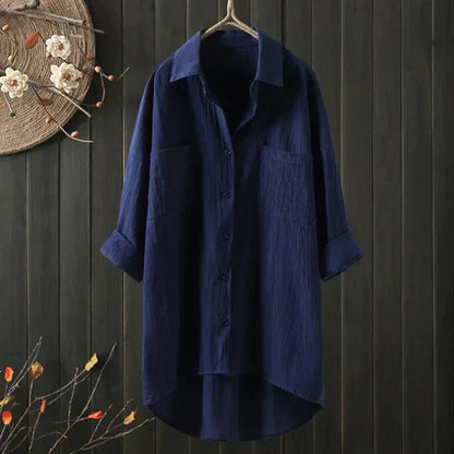 Dunkelblaues, langärmliges Hemd aus Leinen, hängend vor dunklem Holzhintergrund, mit Blumenverzierung. Ideal für lässige Mode und Herbsttrends.