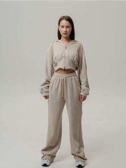 Junge Frau in beigem Jogginganzug mit Kapuzenjacke und Jogginghose, steht vor neutralem Hintergrund. Bequeme Freizeitmode, lässiger Stil.