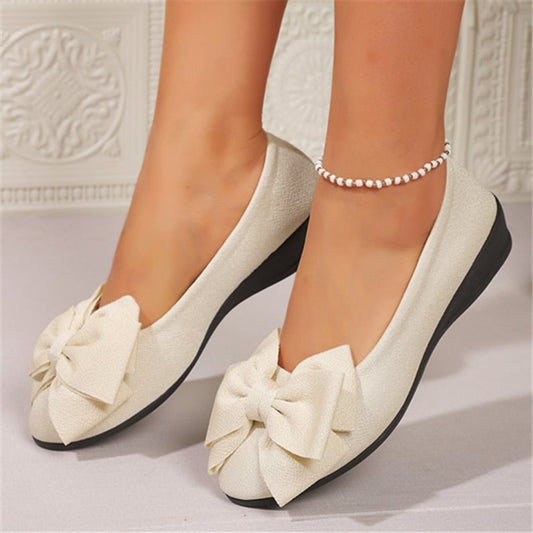 Weiße Ballerinas mit großer Schleife und schwarzer Sohle, getragen von einer Person mit Perlenfußkettchen. Elegante Damenschuhe für besondere Anlässe.