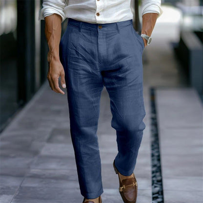 Mann in blauer Leinenhose und weißem Hemd, gehend auf einem Gehweg. Elegante Herrenmode, Sommeroutfit, braune Lederschuhe, stilvoll gekleidet.
