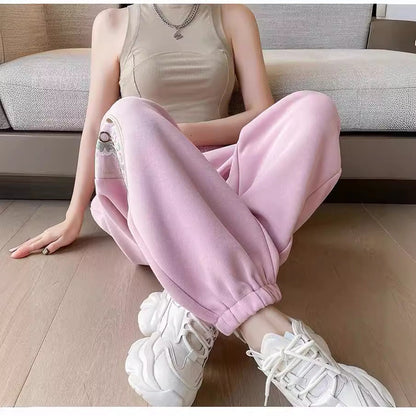 Frau in beiger Weste und rosa Jogginghose sitzt auf Holzboden, trägt weiße Sneaker. Bequeme Freizeitmode, lässiger Stil, moderne Damenbekleidung.