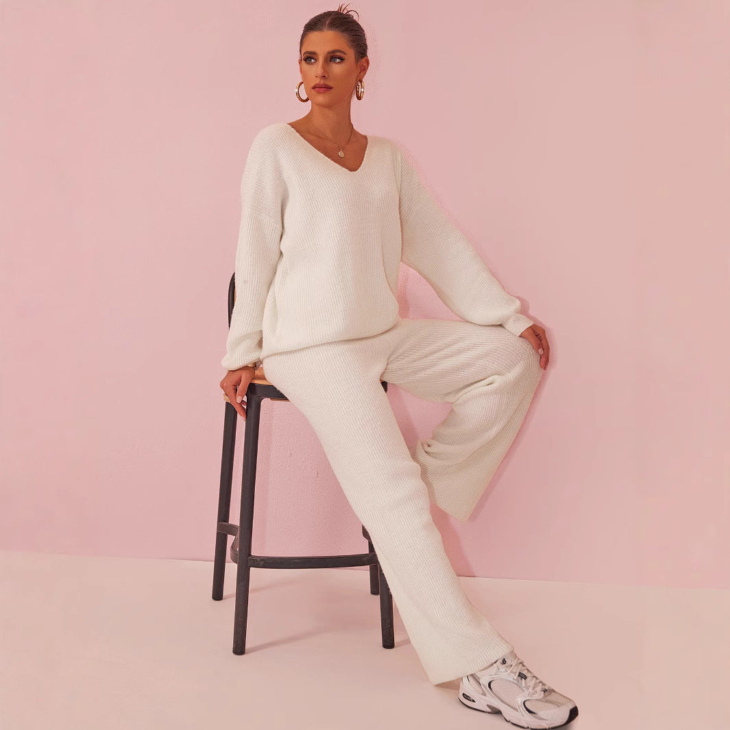 Frau in weißem Strickanzug sitzt auf Stuhl vor rosa Hintergrund, trägt weiße Sneaker. Modetrend, bequeme Damenmode, stilvolles Outfit.