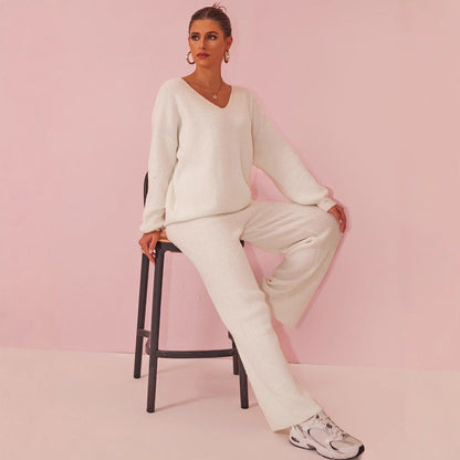 Frau in weißem Strickanzug sitzt auf Stuhl vor rosa Hintergrund, trägt weiße Sneaker. Modetrend, bequeme Damenmode, stilvolles Outfit.