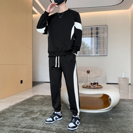 Person in schwarzem Trainingsanzug mit weißen Streifen, steht in modernem Wohnzimmer. Sneaker, Couchtisch und Wandkunst im Hintergrund.