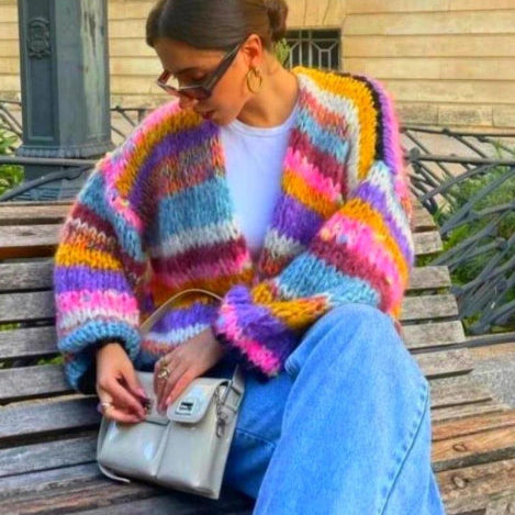 Frau in bunter Strickjacke und blauer Jeans sitzt mit weißer Handtasche. Modetrend, Herbstmode, Streetstyle, lässiger Look.