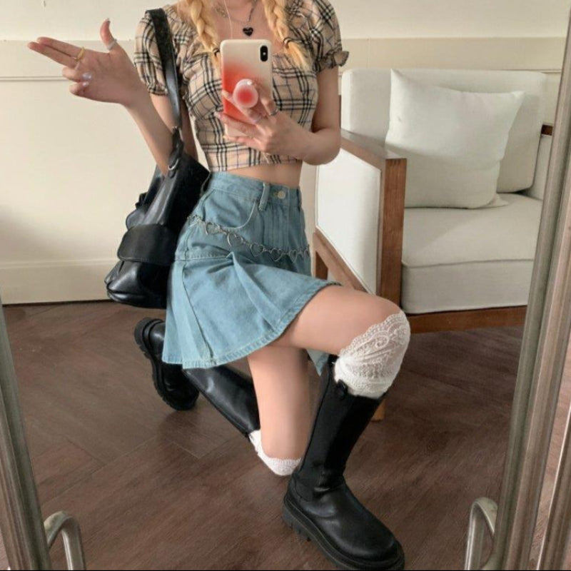 Frau in kariertem Top und Jeansrock, kniet vor Spiegel, trägt schwarze Stiefel und weiße Socken, hält Smartphone, modisches Outfit, Selfie.
